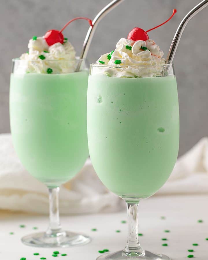 Two shamrock milkshakes topped with whipped topping and cherries in glasses with stainless straws.