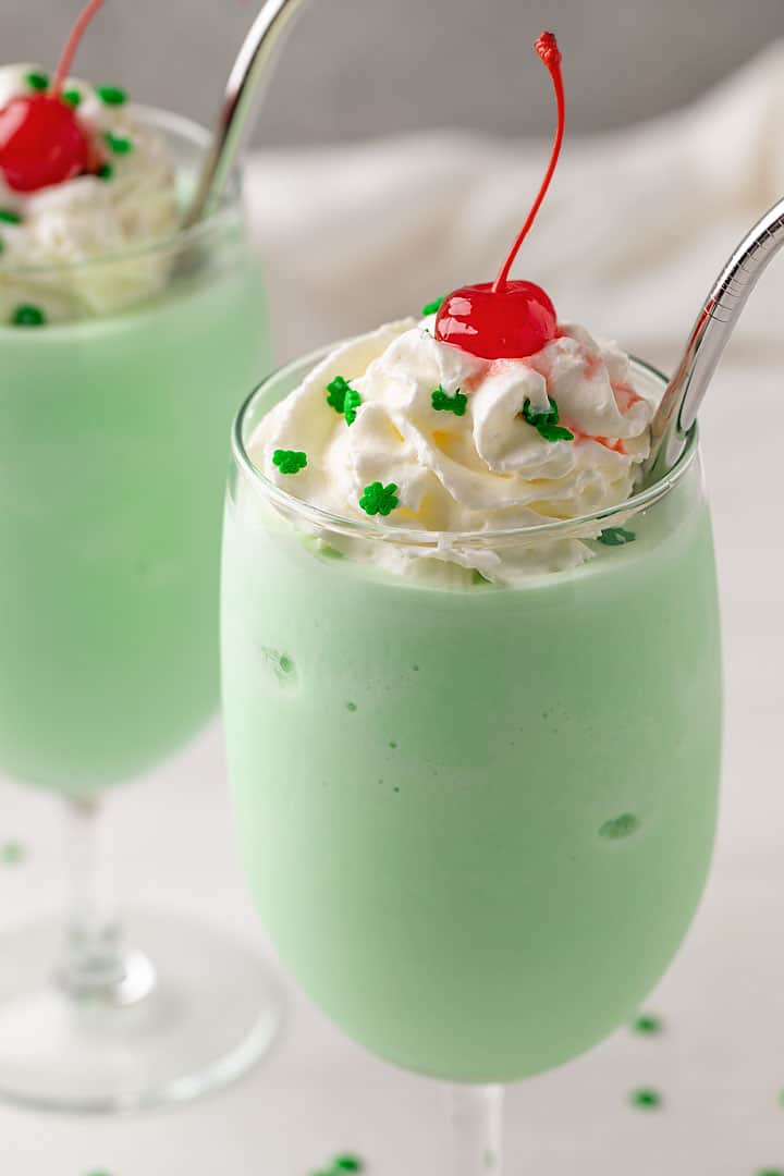A shamrock shake topped with whipped topping, green clover decorator sprinkles and a maraschino cherry.