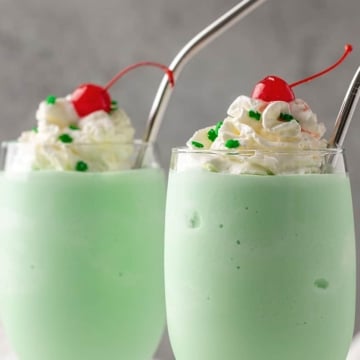 Two shamrock shakes in glasses with stainless straws topped with whipped topping, green sprinkles and maraschino cherries.
