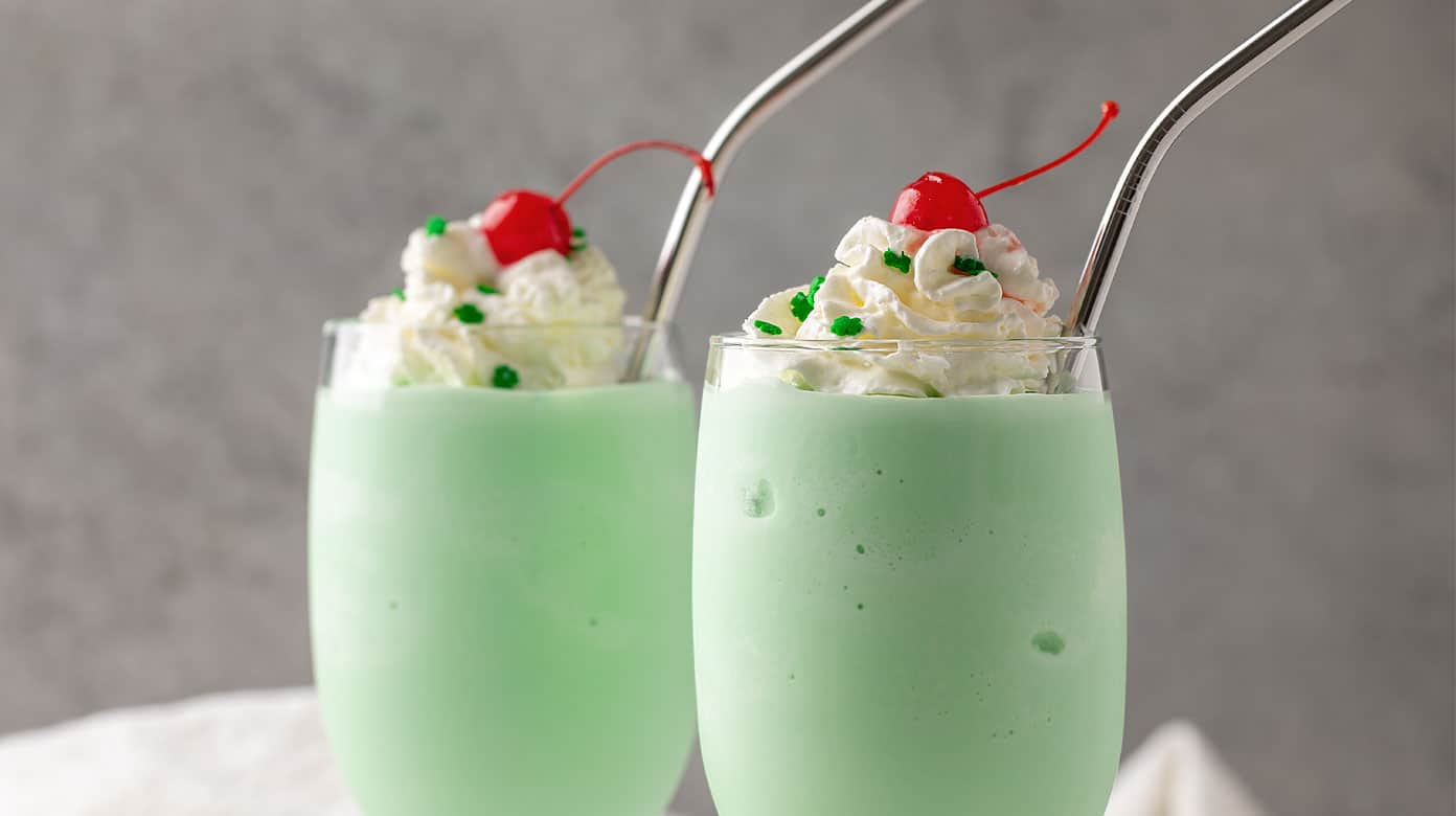 2 shamrock shakes in glasses with stainless straws topped with whipped topping, green sprinkles and maraschino cherries.