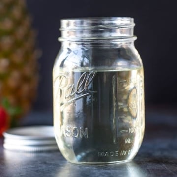A Mason jar filled with simple syrup. A pineapple, lime and strawberry are in the background.