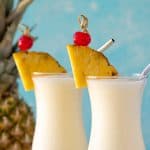 Two pina coladas in glasses with stainless steel straws. A fresh pineapple is in the background.