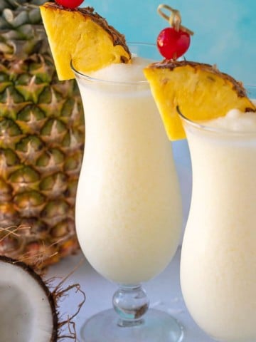 Two pina coladas in hurricane glasses garnished with pineapple wedges and maraschino cherries