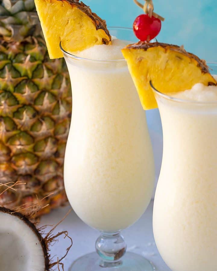 Two pina coladas in hurricane glasses garnished with pineapple wedges and maraschino cherries