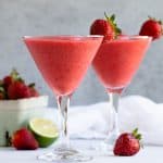 Two daiquiris in glasses with a container of strawberries and a lime in the background.