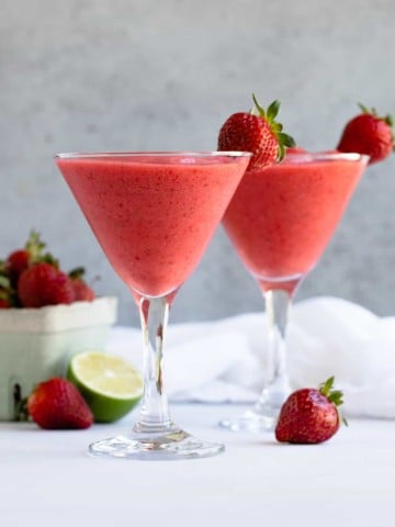 Two daiquiris in glasses with a container of strawberries and a lime in the background.