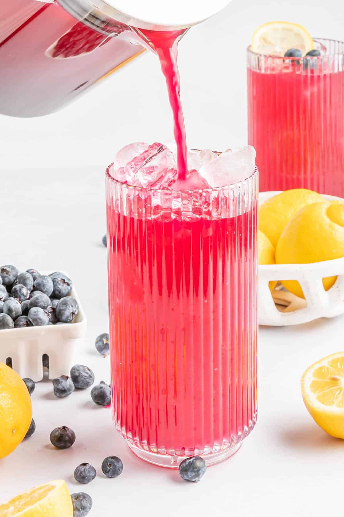 Blueberry lemonade being poured from a pitcher into a glass with ice.