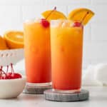 Two sunrise mocktails garnished with an orange slice and a maraschino cherry in highball glasses.