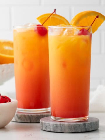 Two sunrise mocktails garnished with an orange slice and a maraschino cherry in highball glasses.